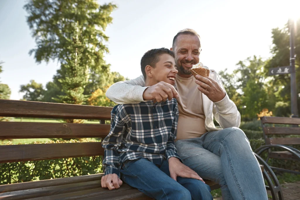 image of happy young disabled boy sitting on park bench with father eating ice cream
